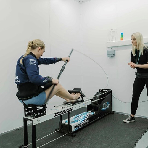 A participant is giving maximum effort on a rowing machine, while a Human Performance Centre specialist measures